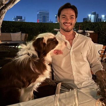 Giovani Urata sentado à mesa de um restaurante ao ar livre, à noite, ao lado do Bruce, um border collie marrom e branco